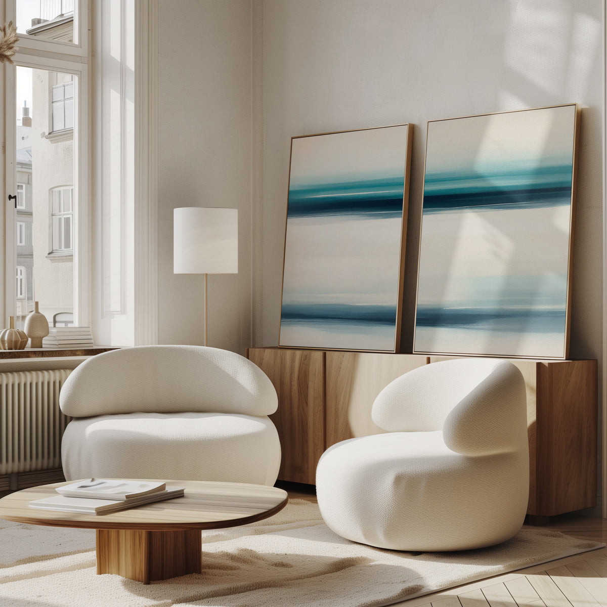 A modern living room with two white rounded armchairs, a low wooden coffee table, and a sideboard. The Ocean Motion - Set Of 2 abstract blue and white paintings add serene sophistication, while large windows bring in natural light.