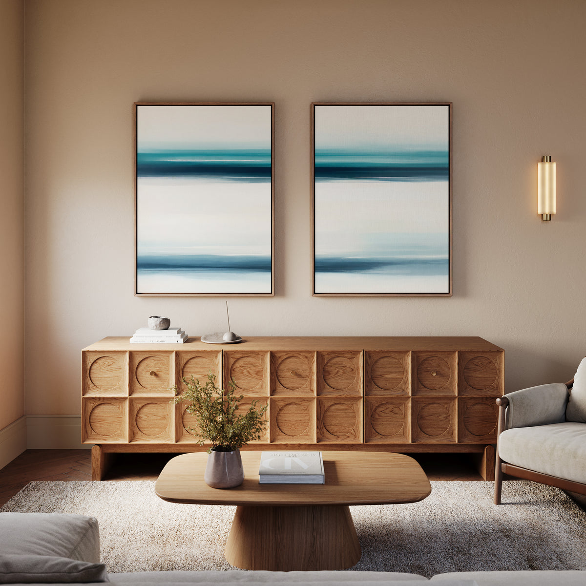 A modern living room exudes serene sophistication with a wooden sideboard, "Ocean Motion - Set Of 2" paintings above it, a beige armchair, wooden coffee table, small plant in a vase, and soft lighting on a neutral wall.