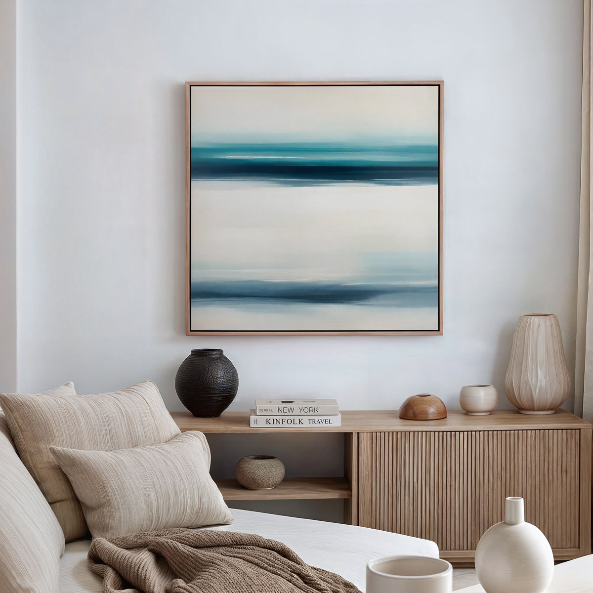 A modern living room features a beige sofa and cushions, a wooden sideboard with books and pottery, and the Ocean Motion Canvas Art - Square above, its blue and white streaks adding serene sophistication to the space.