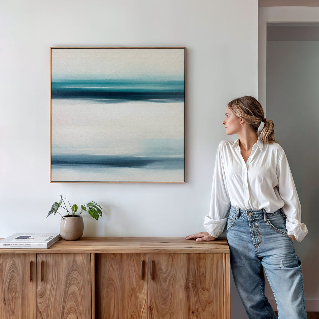 A woman in a white blouse and jeans leans against a wooden cabinet, admiring the Ocean Motion Canvas Art - Square in blue and white above her. A potted plant and book nearby add to the serene, sophisticated atmosphere.