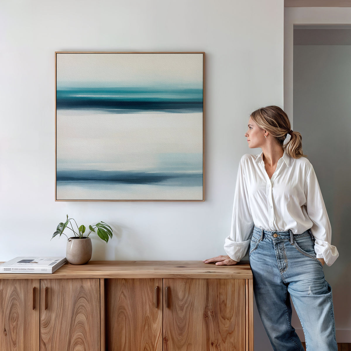 A woman in a white blouse and jeans leans against a wooden cabinet, admiring the Ocean Motion Canvas Art - Square in blue and white above her. A potted plant and book nearby add to the serene, sophisticated atmosphere.