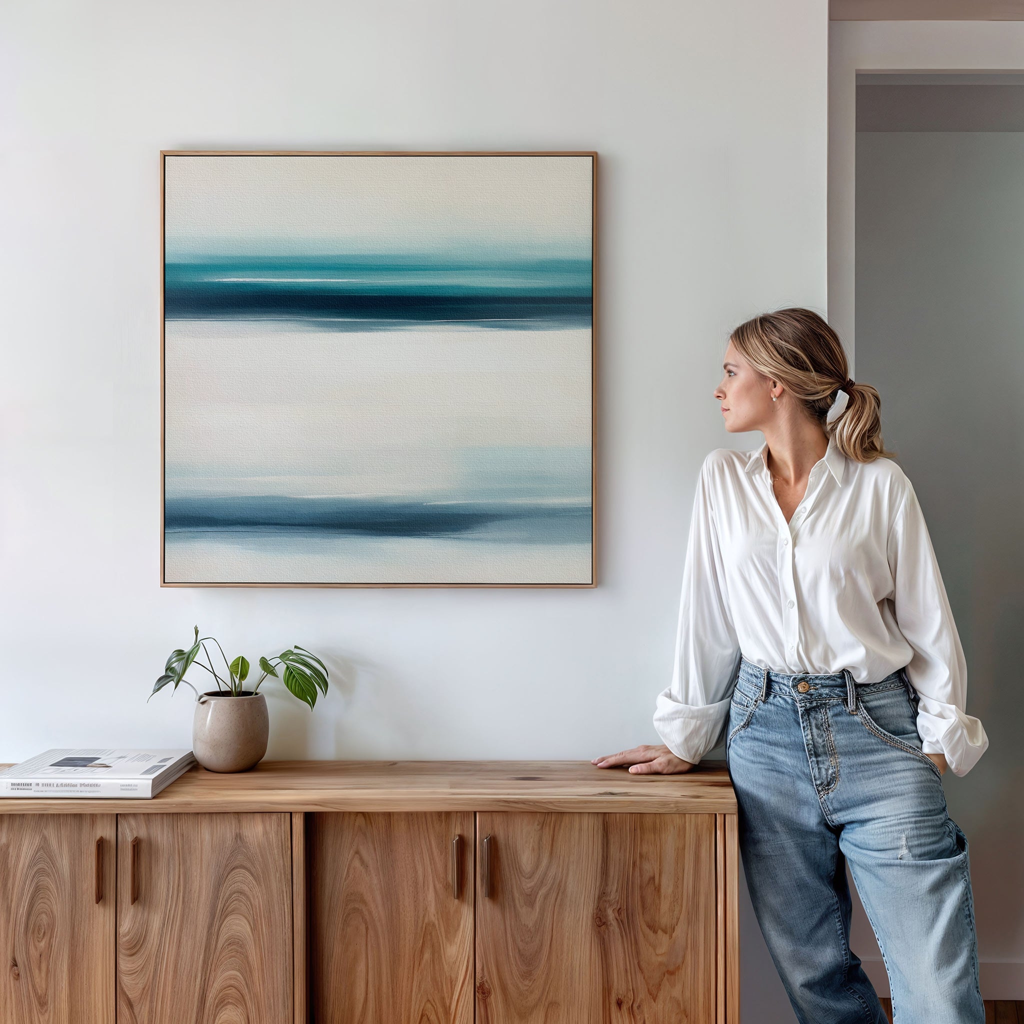 A woman in a white blouse and jeans leans against a wooden cabinet, admiring the Ocean Motion Canvas Art - Square in blue and white above her. A potted plant and book nearby add to the serene, sophisticated atmosphere.