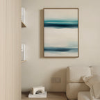 A minimalist living room features a beige sofa, shelves with books and a white decor piece, and the Ocean Motion Canvas Art - Vertical, displaying abstract blue, teal, and white brushstrokes on a cream-colored wall.