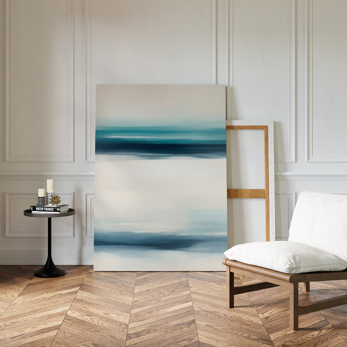 A modern living space with herringbone wood floors, a white cushioned chair, a round black side table with candles, and the Ocean Motion Canvas Art - Vertical in blue and white leaning against a paneled wall exudes serene sophistication.