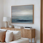 A modern living room features a wooden sideboard with stacked books, a white lamp, and a bowl. Above hangs the Ocean Swells Canvas Art - Horizontal, showcasing calm ocean hues of blue and gray. A white sofa sits in the foreground.