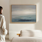 A person in a white outfit stands in a minimal, light-filled room, gazing at the Ocean Swells Canvas Art - Horizontal—a coastal print in blue and gray tones—hanging above a textured cream-colored sofa.