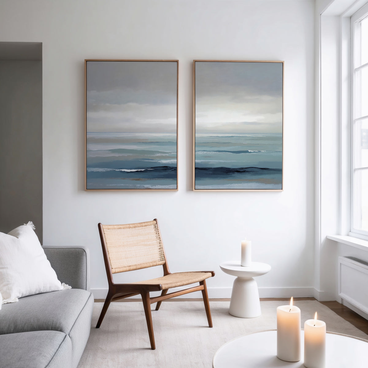 A modern, minimal living room features a rattan chair, gray sofa with a white pillow, two lit candles, and "Ocean Swells - Set Of 2" abstract ocean-themed wall art above a small round table by a large window.