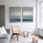 A modern, minimal living room features a rattan chair, gray sofa with a white pillow, two lit candles, and "Ocean Swells - Set Of 2" abstract ocean-themed wall art above a small round table by a large window.