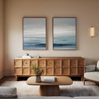 A modern living room with a wooden sideboard, a coffee table with books and a vase, a light sofa, and the "Ocean Swells - Set Of 2" serene abstract landscape wall art displayed above the sideboard.