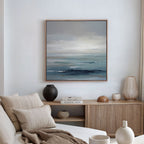 A minimalist living room features a beige sofa, neutral cushions, and a wooden sideboard. Above vases and books hangs the Ocean Swells Canvas Art - Square, infusing Nordic Park calm as natural light streams through the curtain.