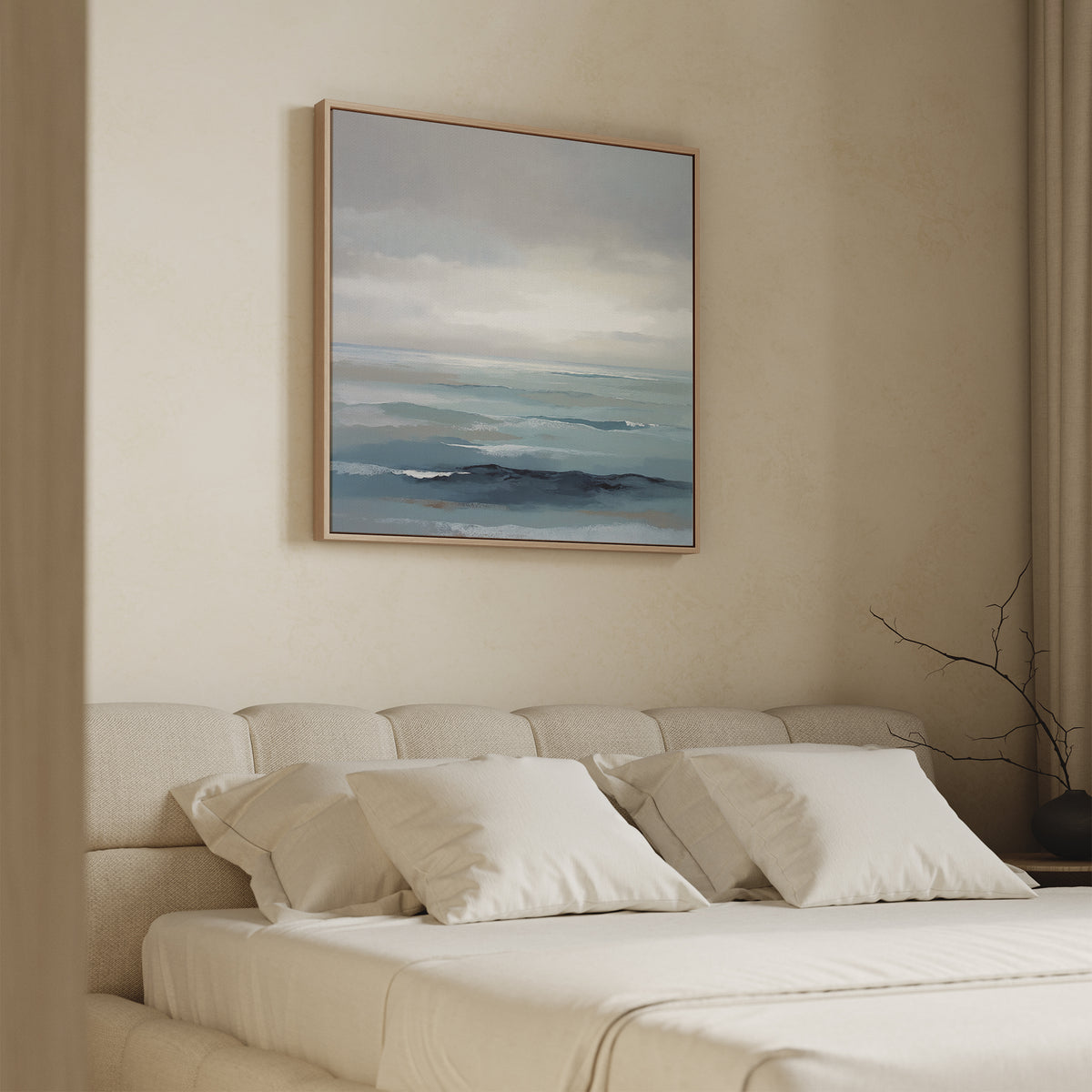 A minimalist bedroom with a beige bed and four pillows features the Ocean Swells Canvas Art - Square, an abstract seascape in soft blue and gray, hung above the headboard. A bare branch in a vase sits on the bedside table.