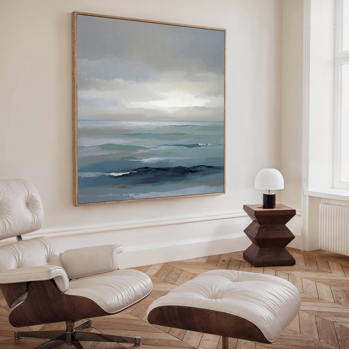 A modern, minimalist room with the Ocean Swells Canvas Art - Square above a white leather lounge chair and ottoman, complemented by a geometric wooden side table, small white lamp, and tall window.