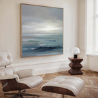 A modern, minimalist room with the Ocean Swells Canvas Art - Square above a white leather lounge chair and ottoman, complemented by a geometric wooden side table, small white lamp, and tall window.