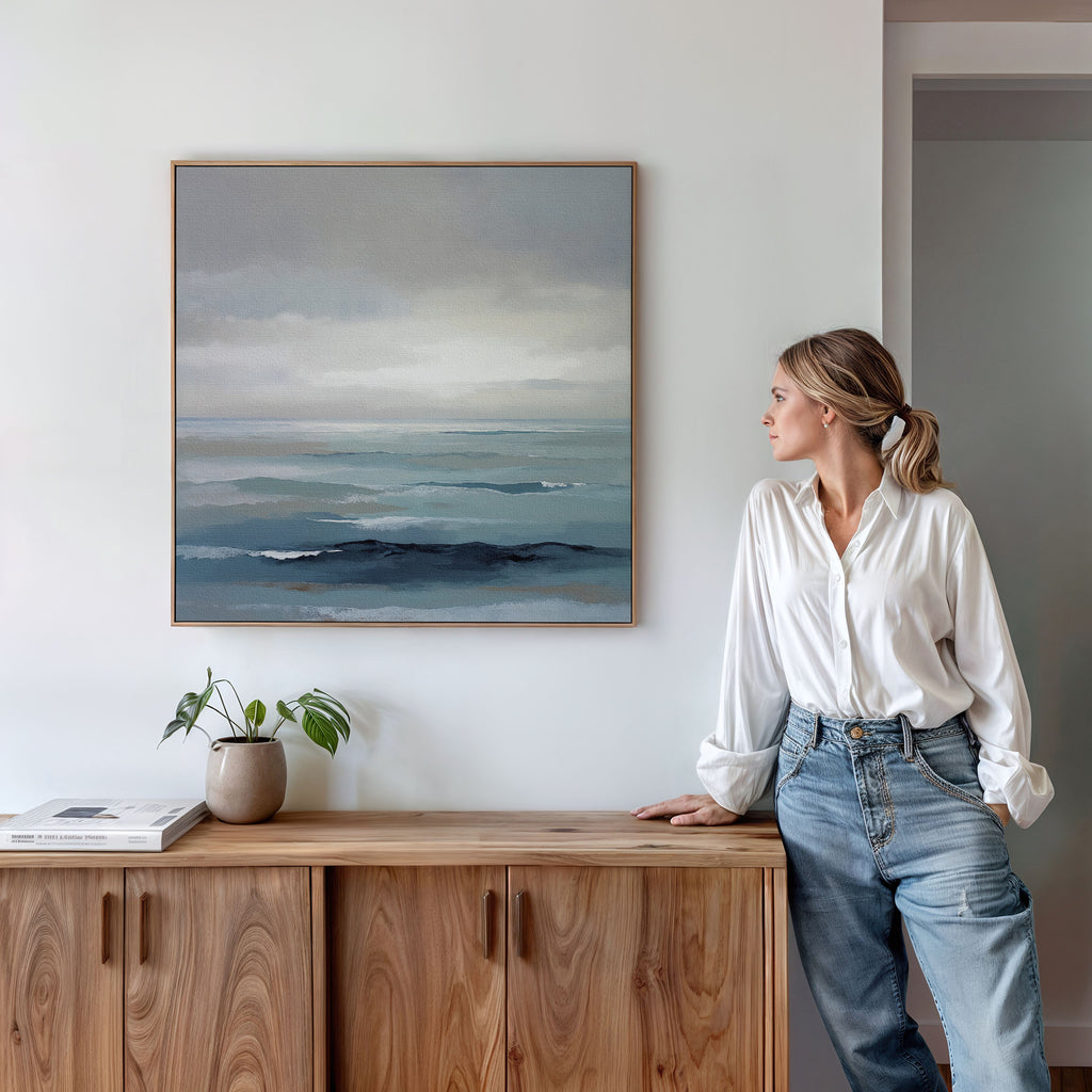 A woman in a white blouse and jeans stands by a wooden sideboard, admiring the Ocean Swells Canvas Art - Square. A small potted plant and stacked books on the sideboard enhance the calm, coastal atmosphere.