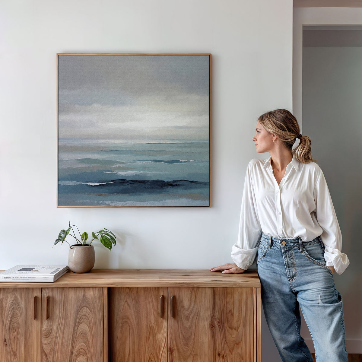 A woman in a white blouse and jeans stands by a wooden sideboard, admiring the Ocean Swells Canvas Art - Square. A small potted plant and stacked books on the sideboard enhance the calm, coastal atmosphere.