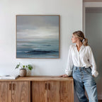 A woman in a white blouse and jeans stands by a wooden sideboard, admiring the Ocean Swells Canvas Art - Square. A small potted plant and stacked books on the sideboard enhance the calm, coastal atmosphere.