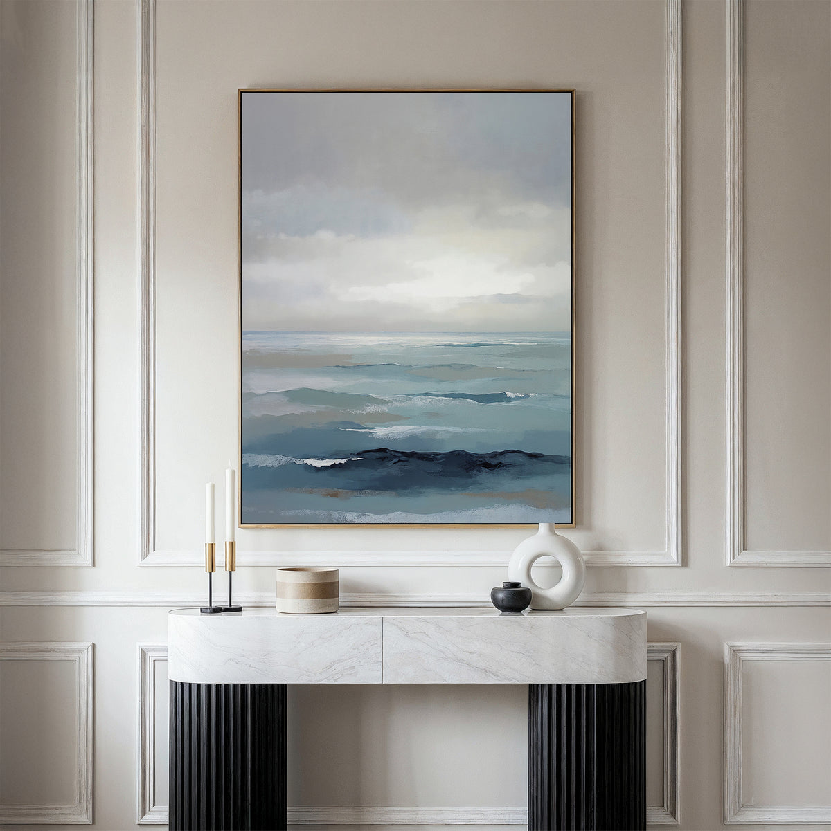 A minimalist room features the Ocean Swells Canvas Art - Vertical in blue and gray tones on a cream-paneled wall above a marble console with decor accents, adding a tranquil, serene touch to the space.