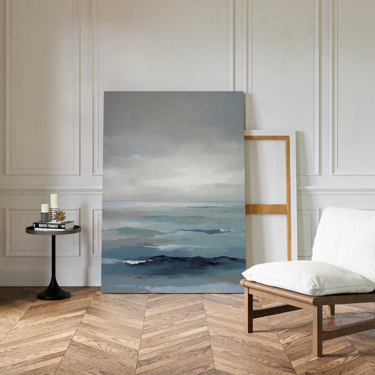 A minimalist room with chevron wood floors, a white cushioned chair, and a black side table with candles. The Ocean Swells Canvas Art - Vertical leans against the paneled wall beside an empty wooden frame.