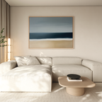 A modern living room features a light beige sectional sofa, a round wooden coffee table, and the Ocean Edge Canvas Art – Horizontal above. Soft natural light streams in from the left, with a green plant partially visible.