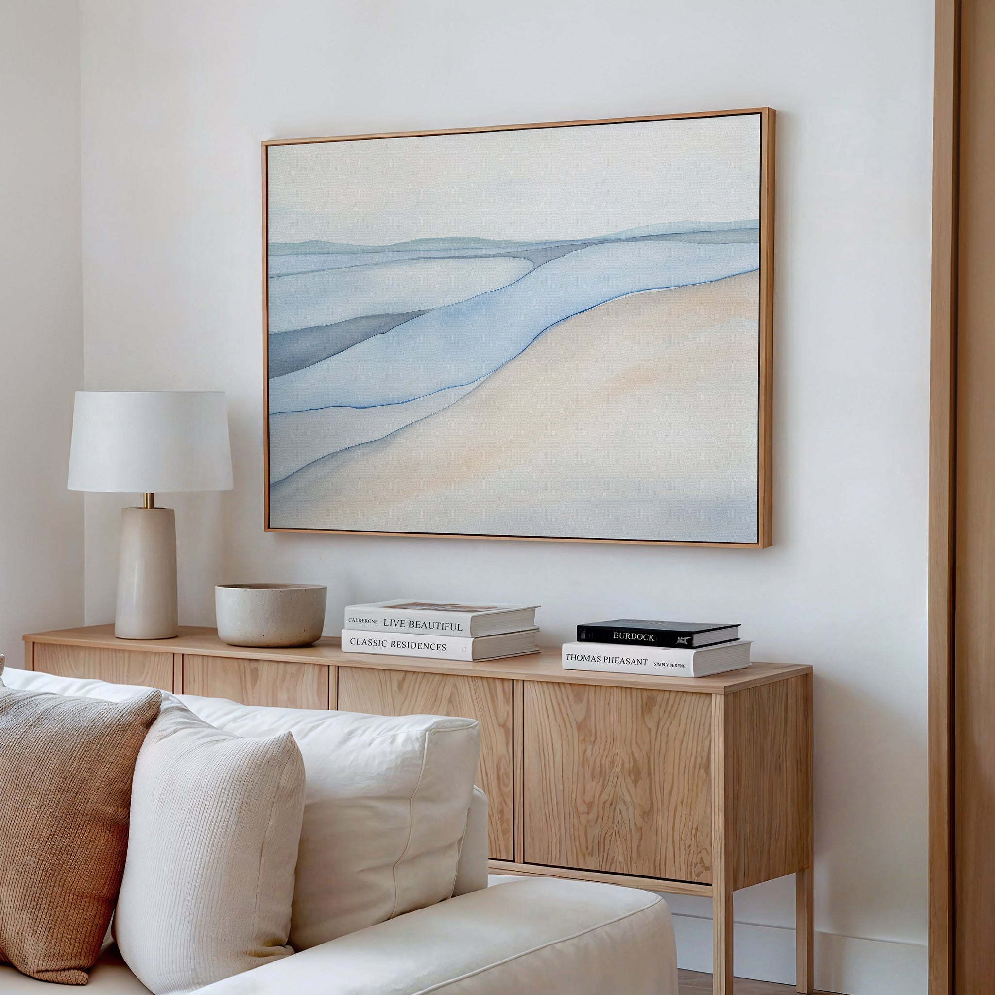A minimal, modern living room features a light wood sideboard, neutral décor, stacked books, a white lamp, and calming artwork—a large Pale Currents Canvas Art - Horizontal in soft blue, beige, and cream tones—hung above.