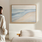 A person in a white robe stands by a beige sofa, admiring the Pale Currents Canvas Art - Horizontal, a soothing abstract piece of soft blue and sandy tones, displayed on a light wall in a minimalist room.