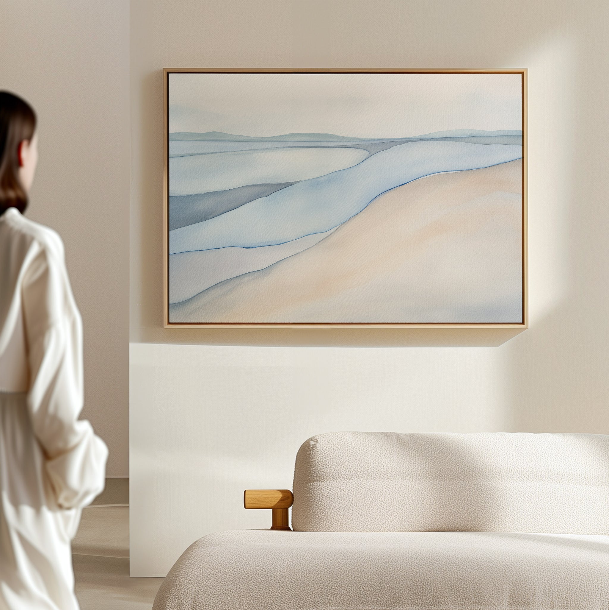 A person in a white robe stands by a beige sofa, admiring the Pale Currents Canvas Art - Horizontal, a soothing abstract piece of soft blue and sandy tones, displayed on a light wall in a minimalist room.