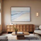 A minimalist living room features a wooden coffee table, textured sideboard, light sofa, neutral rug, and calming artwork—a Pale Currents Canvas Art - Horizontal with blue and beige tones on a beige wall. A wall sconce offers soft lighting.
