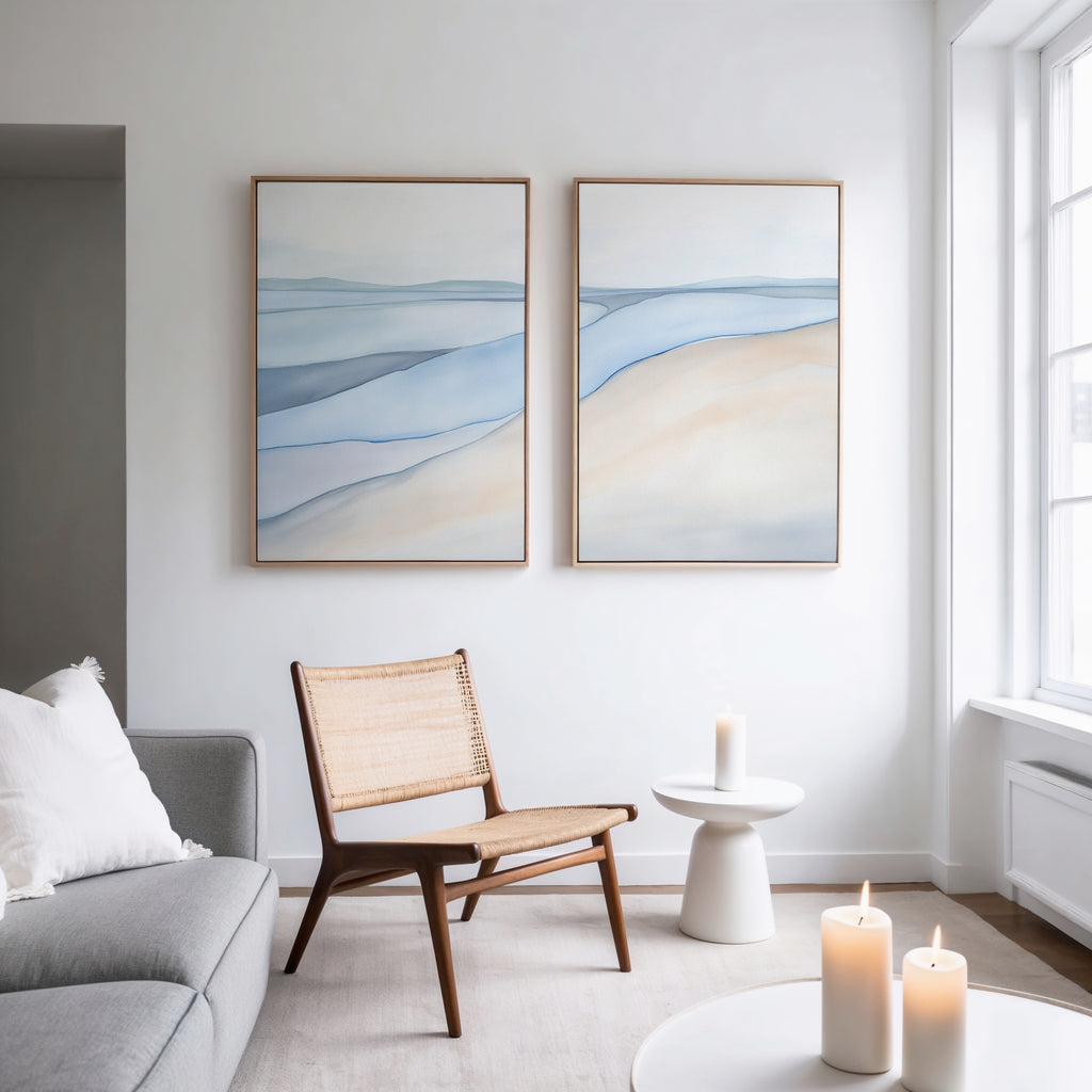A modern, minimalist living room features a woven chair, small white table, lit candles, and calming artwork—Pale Currents - Set of 2 abstract blue and beige canvas prints on a white wall beside a large window.