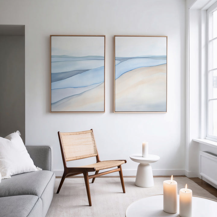 A modern, minimalist living room features a woven chair, small white table, lit candles, and calming artwork—Pale Currents - Set of 2 abstract blue and beige canvas prints on a white wall beside a large window.