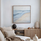 A minimalist living room with a beige sofa, neutral cushions, wooden sideboard, vases, stacked books, and the Pale Currents Canvas Art - Square that brings Nordic Park-inspired calm to the space.
