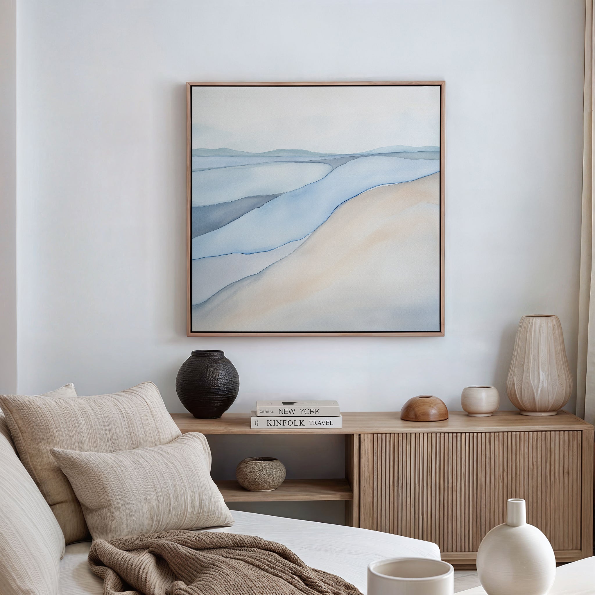 A minimalist living room with a beige sofa, neutral cushions, wooden sideboard, vases, stacked books, and the Pale Currents Canvas Art - Square that brings Nordic Park-inspired calm to the space.