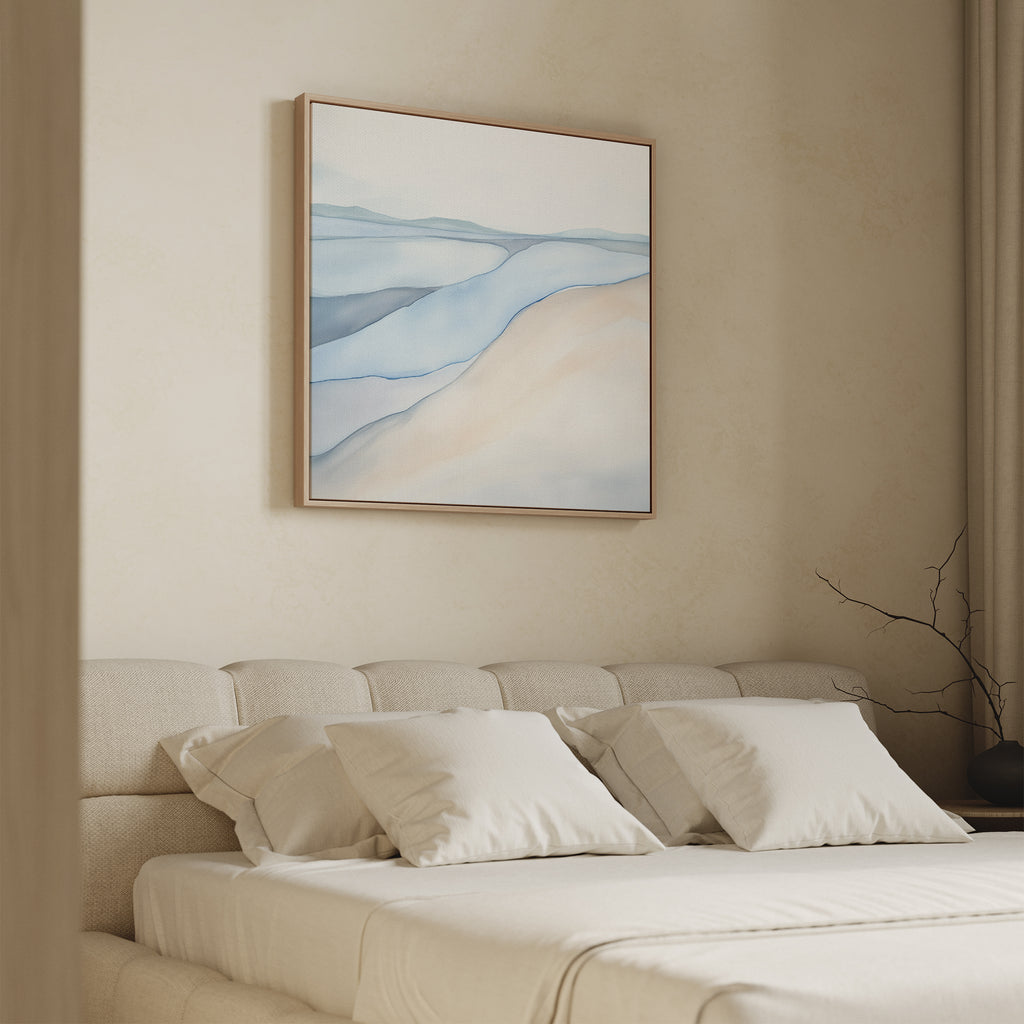 A minimalistic bedroom features a beige upholstered bed with white pillows and bedding. Above the bed hangs the Pale Currents Canvas Art - Square, an abstract print in soft blue and beige tones. A bare branch decorates the right nightstand.