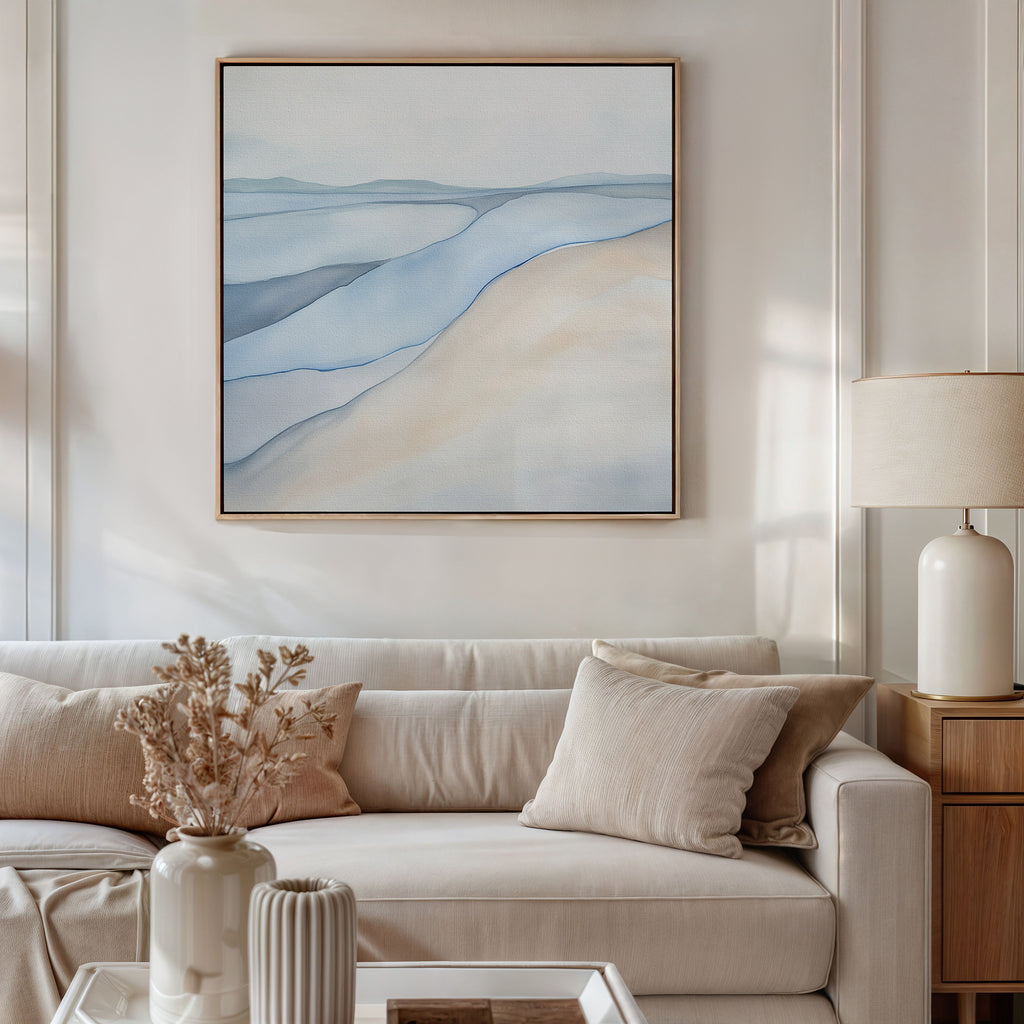 A modern living room with a beige sofa, neutral cushions, a dried floral arrangement in a vase, a table lamp, and the Pale Currents Canvas Art - Square—a large abstract print in soft blue and beige tones—hanging on the wall.