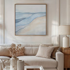 A modern living room with a beige sofa, neutral cushions, a dried floral arrangement in a vase, a table lamp, and the Pale Currents Canvas Art - Square—a large abstract print in soft blue and beige tones—hanging on the wall.