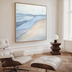 A modern, minimalist living room featuring a white leather lounge chair and ottoman, a geometric wooden side table with a dome lamp, and the Pale Currents Canvas Art - Square in soft blue and beige tones on the wall.