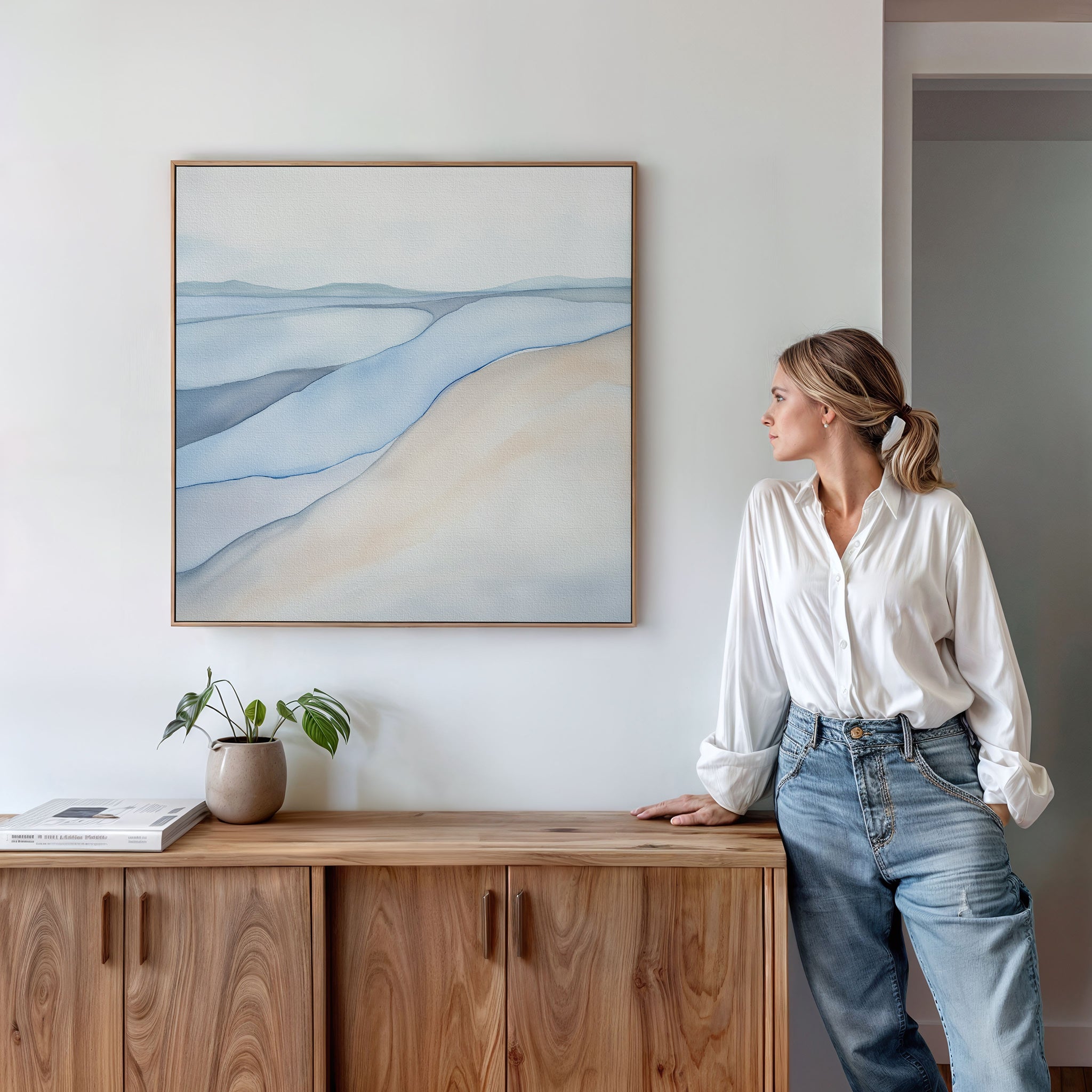 A woman in a white blouse and jeans leans against a wooden cabinet, gazing at the Pale Currents Canvas Art - Square. A potted plant and books rest on the cabinet in this modern, bright room.