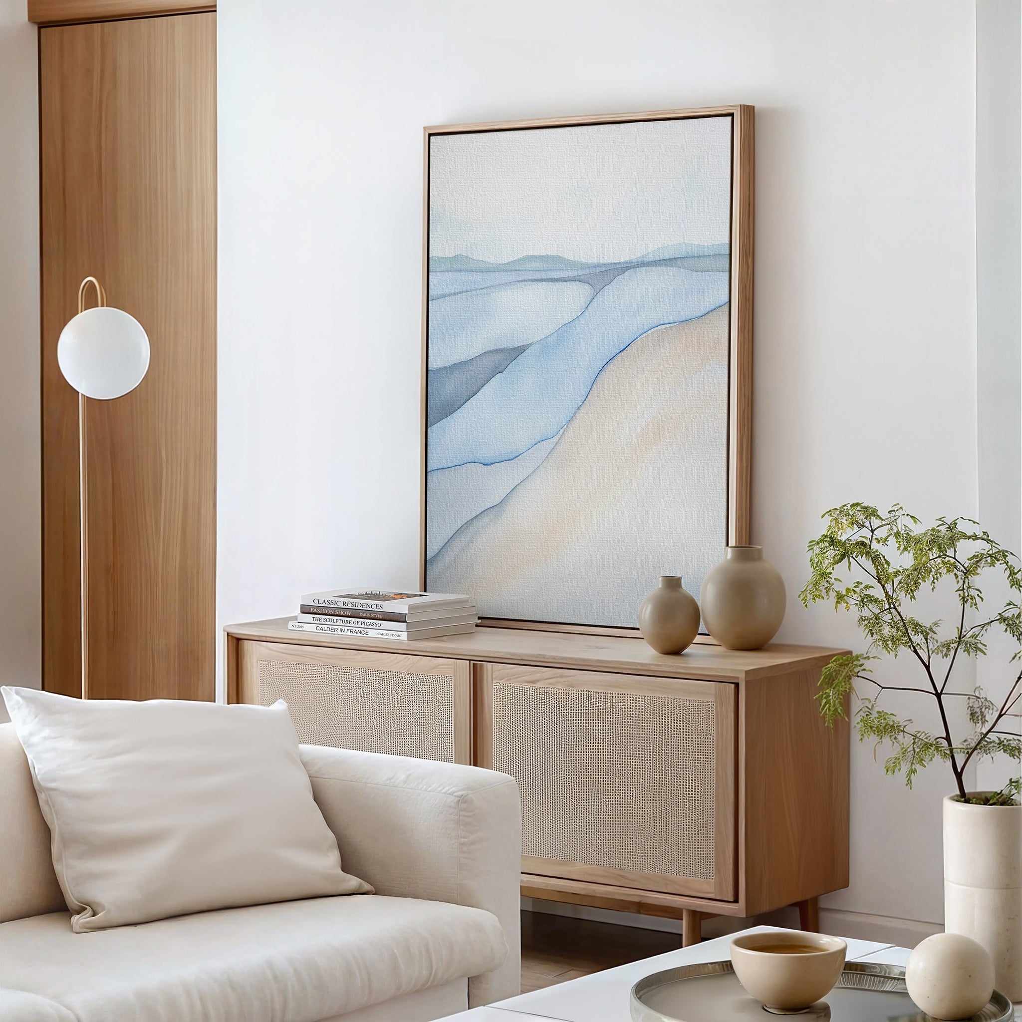 A modern living room with neutral tones features the Pale Currents Canvas Art - Vertical, showcasing calming blue and beige waves above a light wood sideboard with ceramics, books, and a small leafy plant.