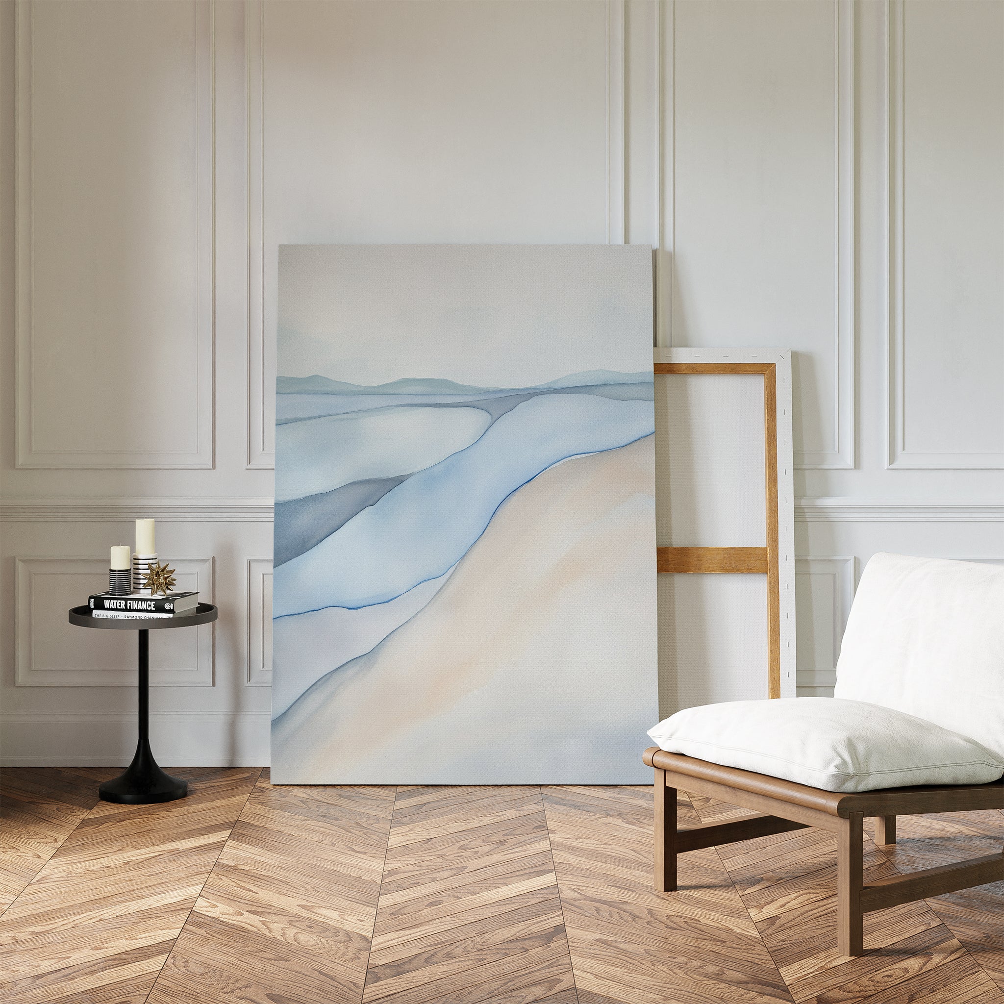 The Pale Currents Canvas Art - Vertical, featuring soft blue and beige tones, leans against a white paneled wall beside an empty wooden frame. A cushioned chair and candles nearby add calming Nordic Park-inspired vibes to the space.