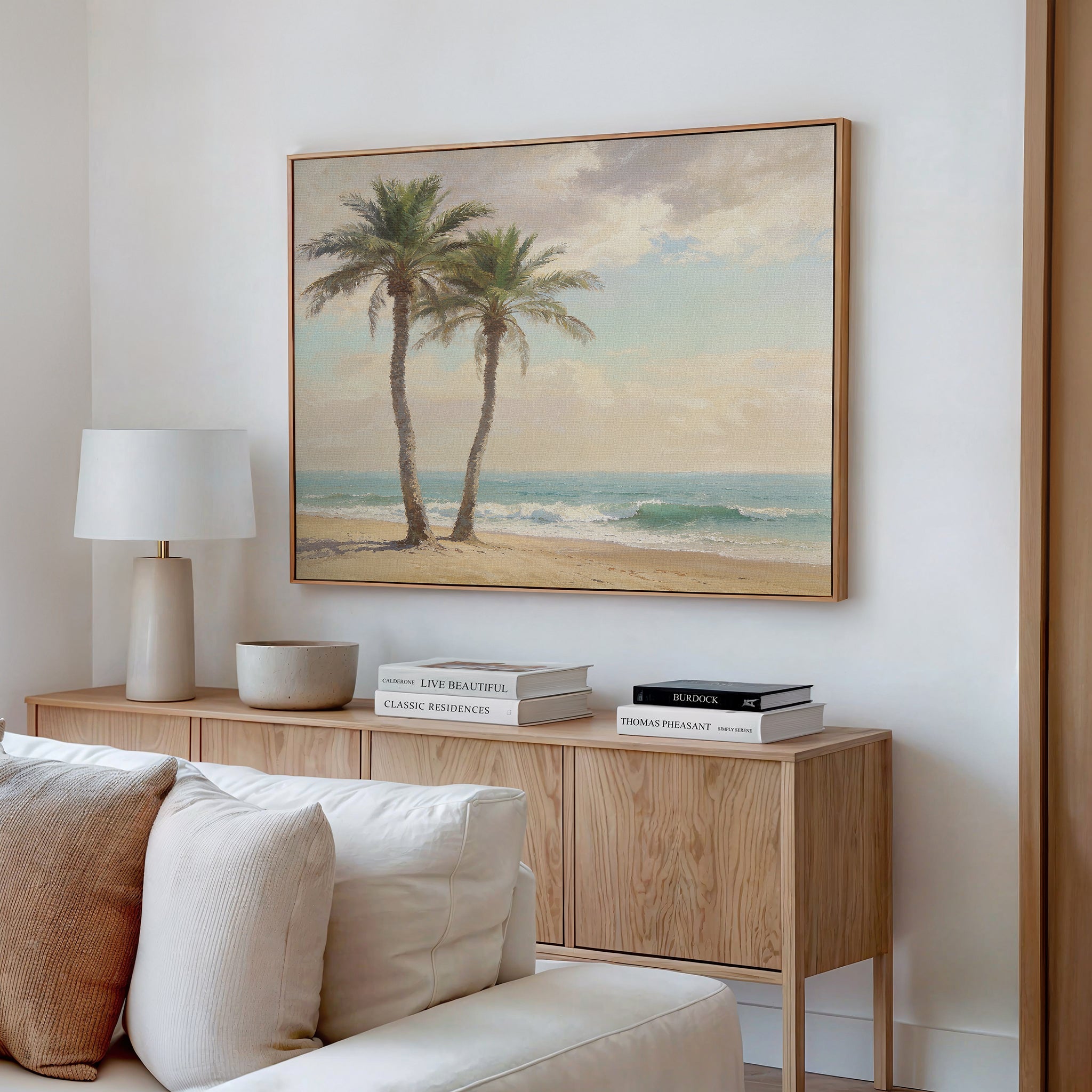 A cozy, modern living room with a wooden sideboard, beige sofa, white lamp, decorative bowl, and stacked books. Above the sideboard hangs Paradise Found Canvas Art - Horizontal—two palm trees on a sandy beach with ocean waves.