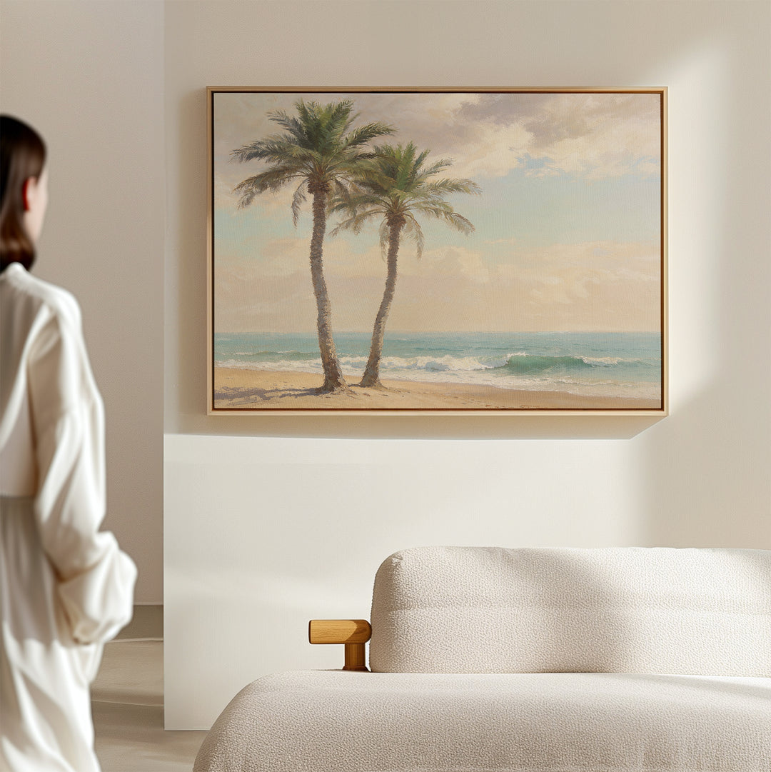 A person in a white shirt stands in a minimal, sunlit room with a beige sofa and the Paradise Found Canvas Art - Horizontal, featuring two palm trees on a sandy shore, hanging on the wall.