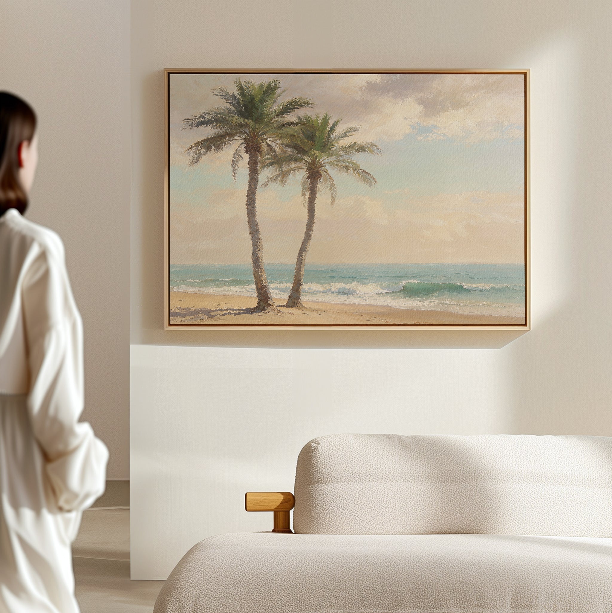A person in a white shirt stands in a minimal, sunlit room with a beige sofa and the Paradise Found Canvas Art - Horizontal, featuring two palm trees on a sandy shore, hanging on the wall.