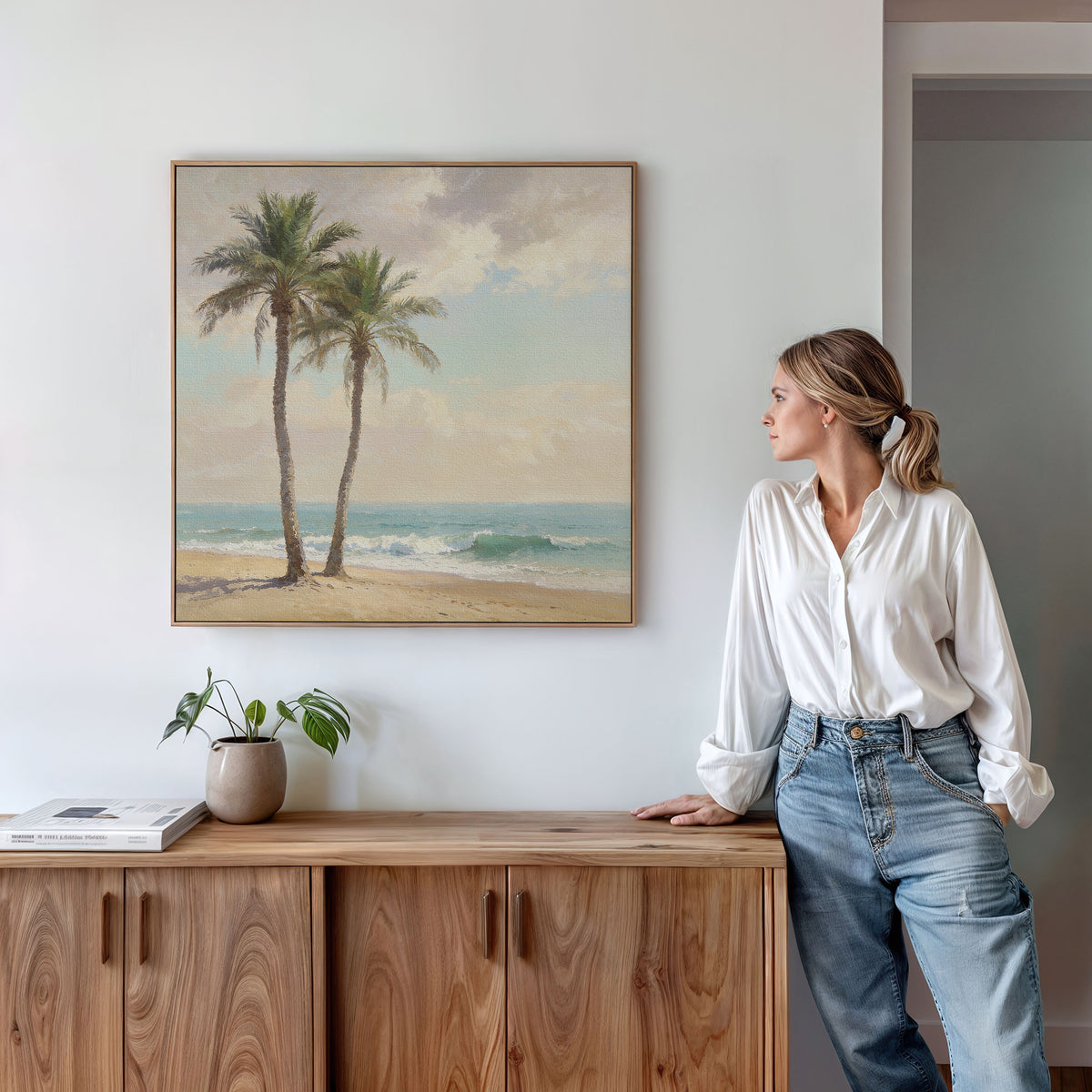 A woman in a white blouse and jeans stands by a wooden cabinet, admiring the Paradise Found Canvas Art - Square featuring two palm trees above her. On the cabinet sits a potted plant and a closed book.