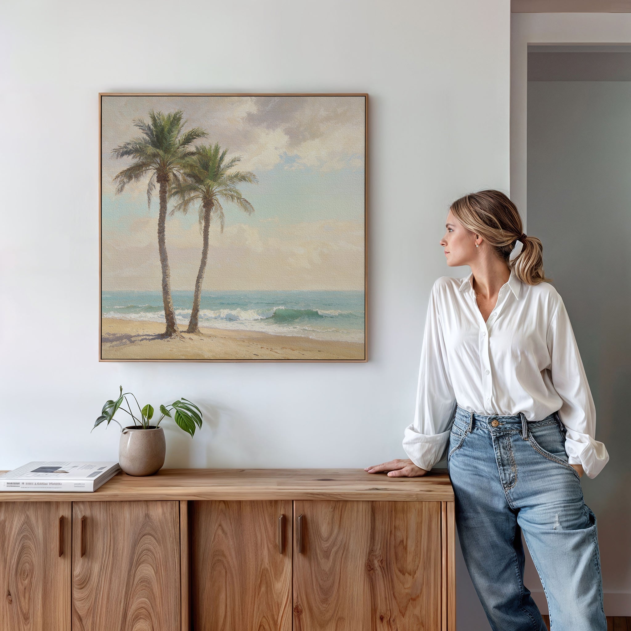 A woman in a white blouse and jeans stands by a wooden cabinet, admiring the Paradise Found Canvas Art - Square featuring two palm trees above her. On the cabinet sits a potted plant and a closed book.