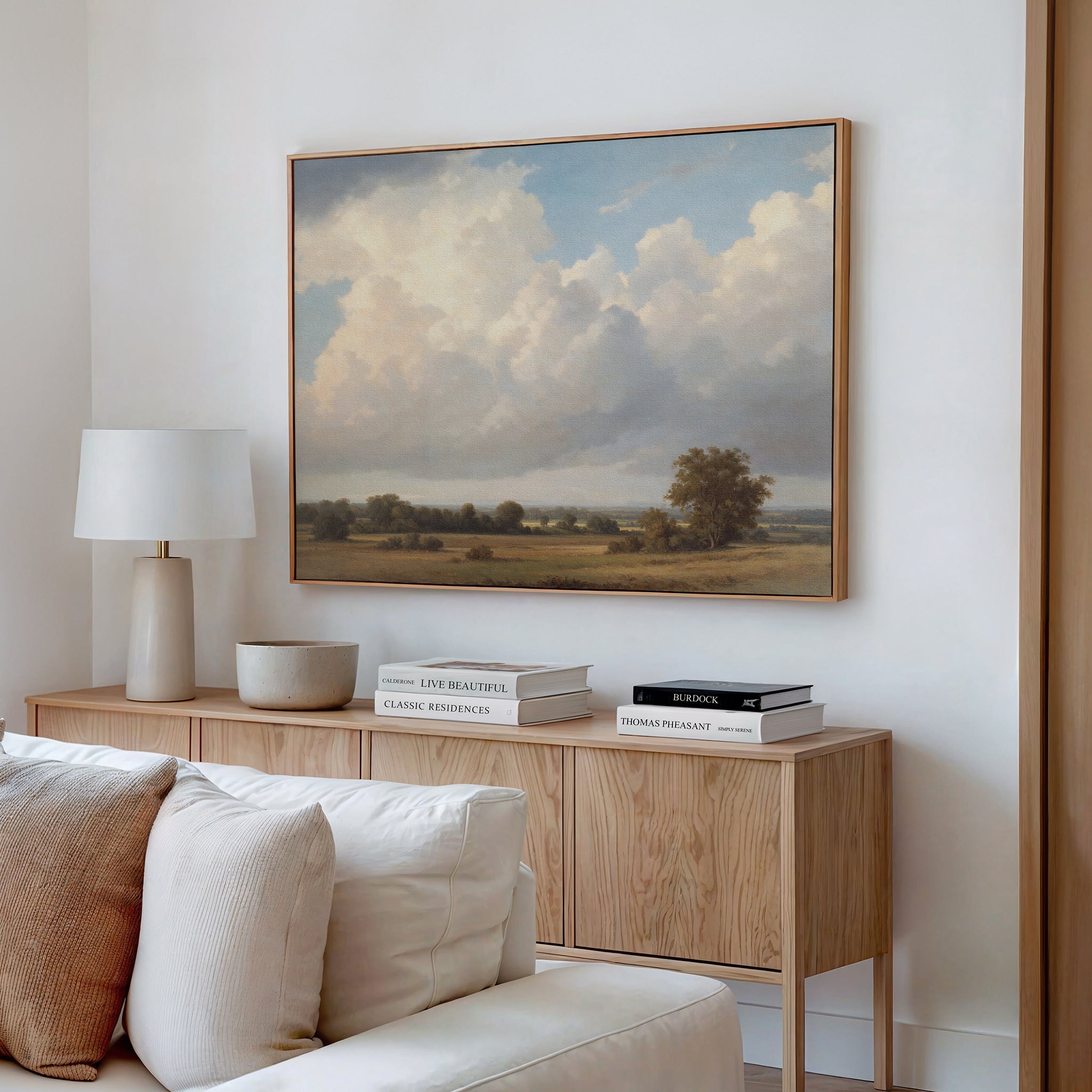 A cozy living room showcases a wooden sideboard with books, lamp, and bowl, topped by the Pastoral Grandeur Canvas Art - Horizontal depicting a cloudy sky and trees above a cream-colored sofa on a white wall.
