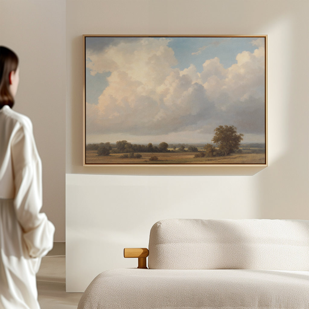 A person in a white shirt admires the 'Pastoral Grandeur Canvas Art - Horizontal' depicting a cloudy sky and open fields, displayed above a light sofa in this minimalist, Nordic Park-inspired room.