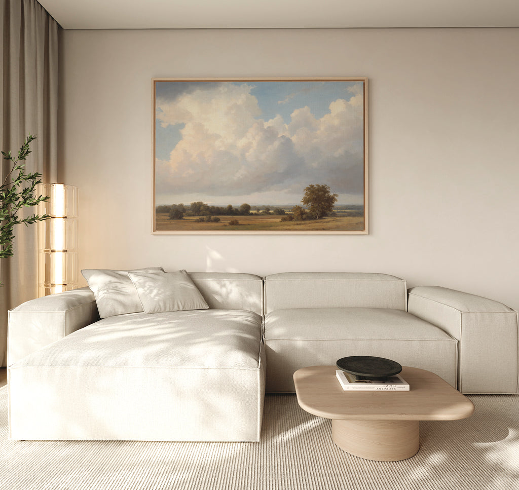 A modern living room with a light beige sectional sofa, wooden coffee table, potted plant, and the Pastoral Grandeur Canvas Art - Horizontal depicting a cloudy sky and trees hanging on a neutral wall.