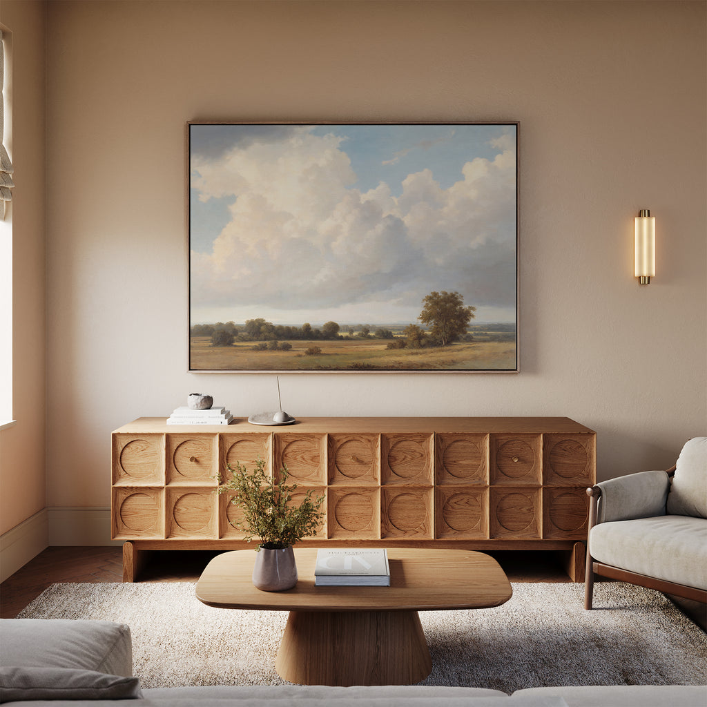 A serene living room with a beige sofa, wooden coffee table, and a geometric-patterned sideboard. The Pastoral Grandeur Canvas Art - Horizontal hangs above the sideboard, complemented by a small wall sconce providing soft lighting.