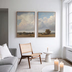 A modern living room with a light gray sofa, woven chair, and round white side table features "Pastoral Grandeur - Set of 2" wall art—large countryside landscapes with clouds and fields—hanging above. Sunlight streams through tall windows.