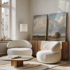 A bright, modern living room with two white rounded armchairs, a wooden coffee table, a floor lamp, and the Pastoral Grandeur - Set of 2 wall art pieces resting on a wooden sideboard against the wall.