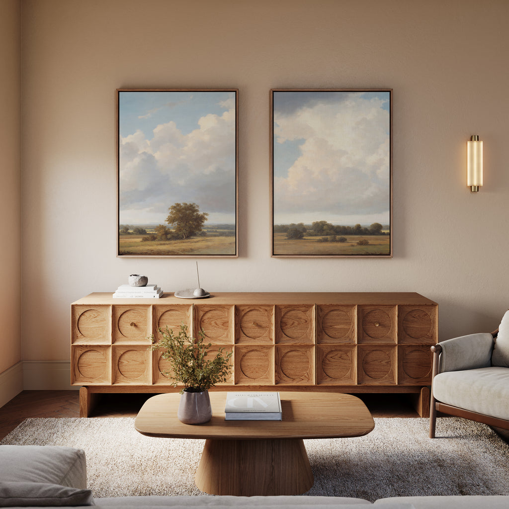 A cozy living room with a wooden sideboard, two "Pastoral Grandeur - Set of 2" wall art pieces above it, a wooden coffee table with a book and flowers, a light gray armchair, and a wall-mounted light fixture.